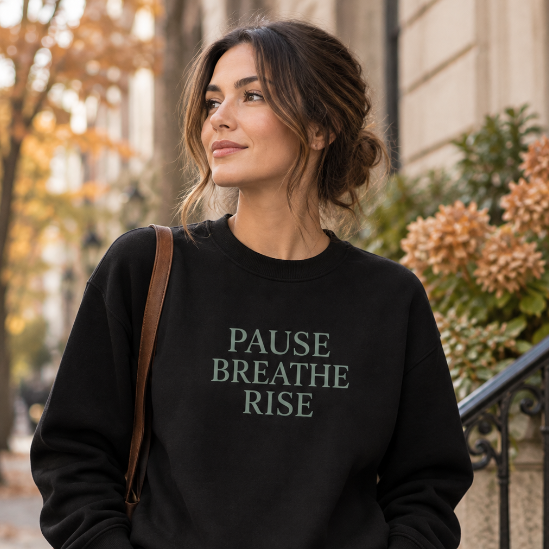 A woman wearing a black crewneck sweatshirt with the text 'PAUSE BREATHE RISE' printed in muted green on the front, photographed outdoors on a city street.