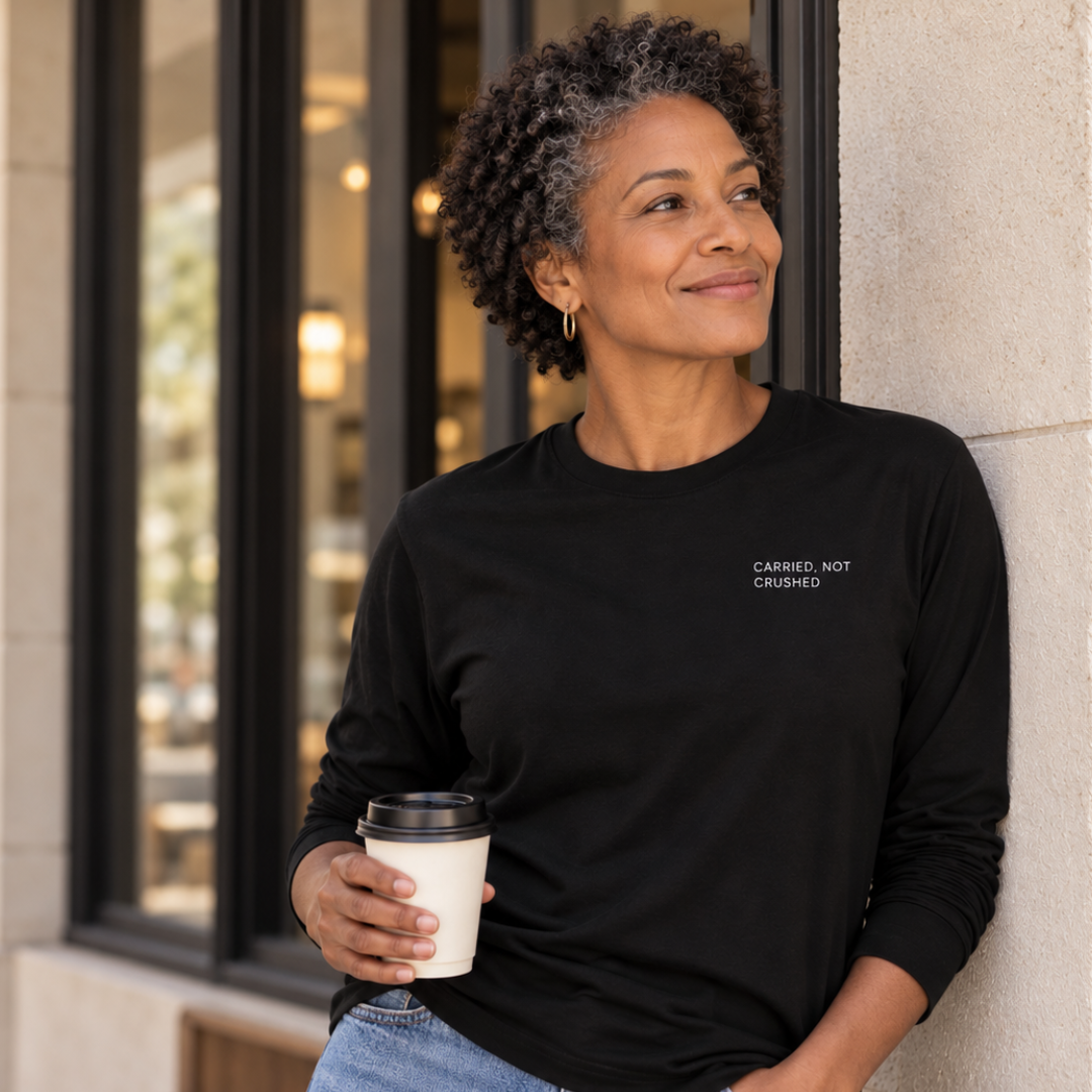 Woman wearing a black long sleeve tee with CARRIED, NOT CRUSHED on the upper left chest, holding a to go coffee cup and learning against a modern building.
