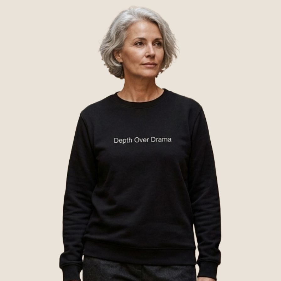 A woman with short gray hair wearing a black crewneck sweatshirt with the white printed text 'Depth Over Drama' on the chest, set against a solid light beige background.