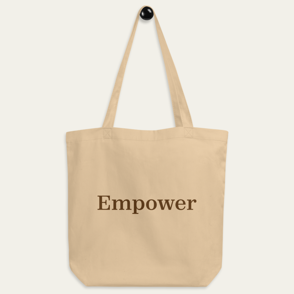 A beige tote bag hanging on a peg. The word "Empower" in brown is centered on the tote.