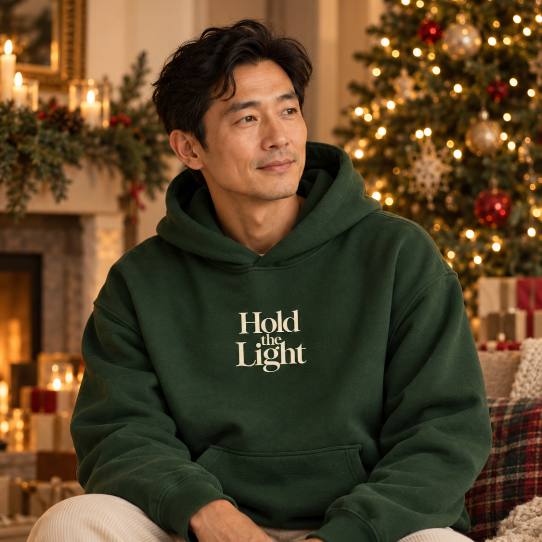 Man wearing a forest green Hold the Light hoodie with a seasonal background with a christmas tree, candles, and fireplace.