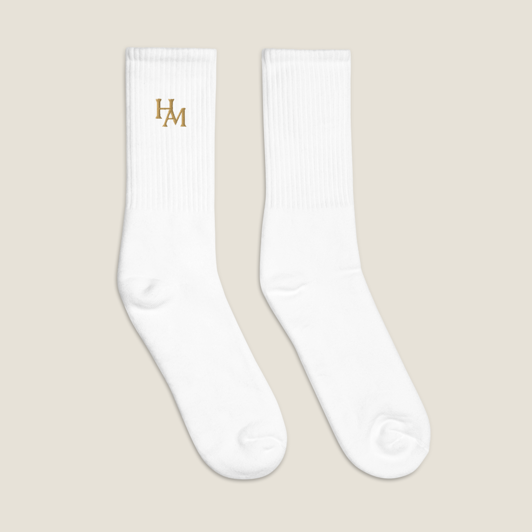 A pair of white ribbed crew socks on a light beige background, featuring a gold embroidered 'HAM' logo on the left sock's cuff.