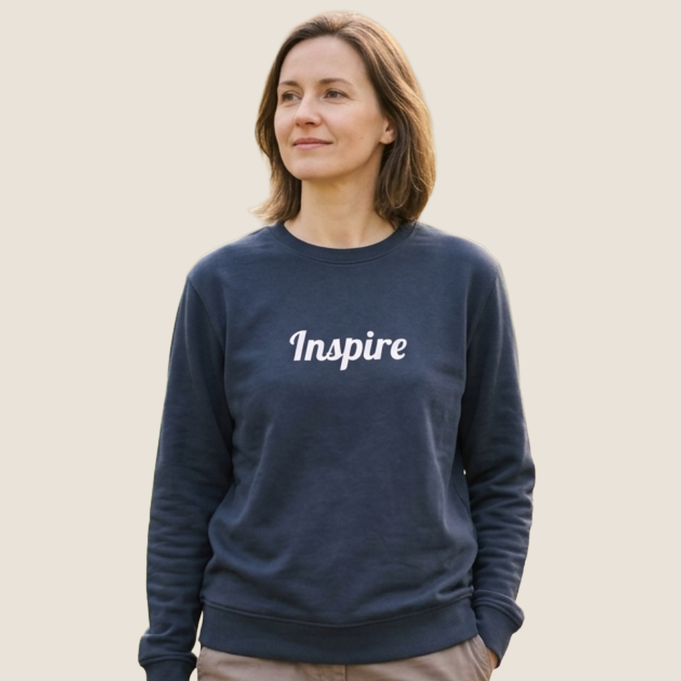 A woman wearing a navy blue crewneck sweatshirt with the word 'Inspire' printed in white script across the chest, paired with beige pants, set against a solid light beige background.