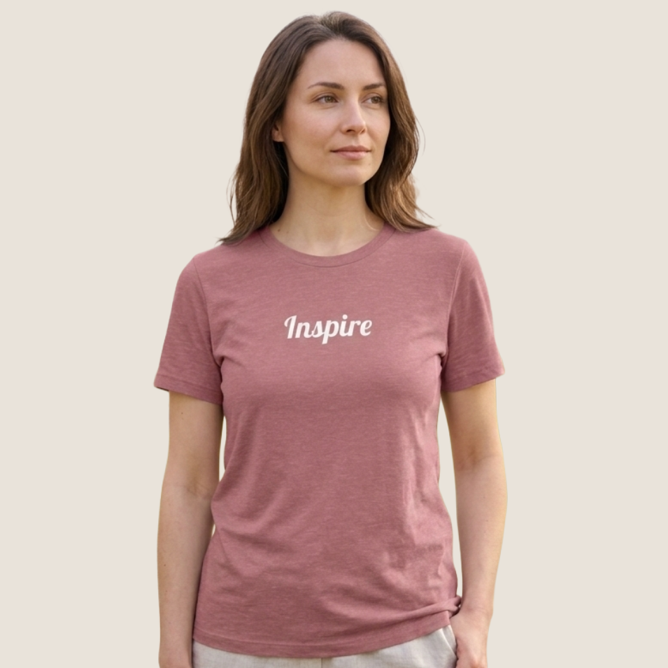 A woman wearing a heathered dusty rose short-sleeve t-shirt with the word 'Inspire' printed in white script across the chest, set against a solid light beige background.