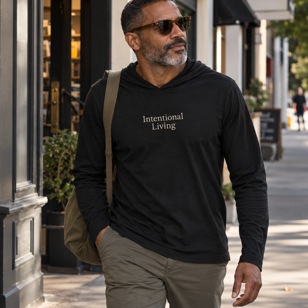 Man walking down a city street wearing a black hooded long sleeve tee with Intentional Living on the chest along with brown pants and carrying a shoulder bag.
