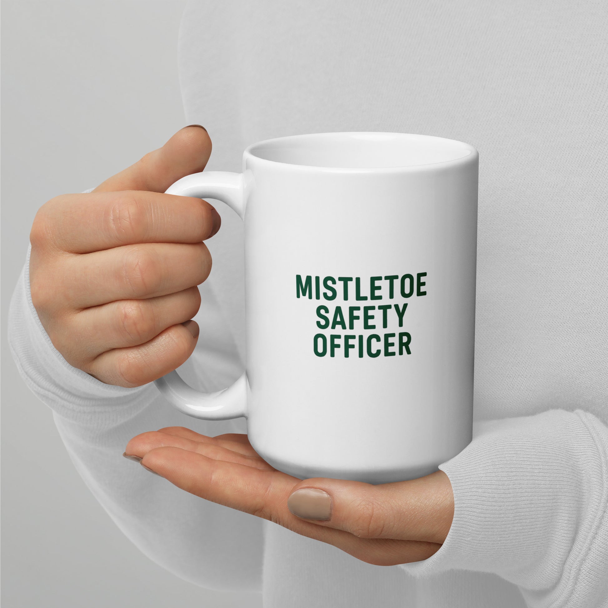 White mug with 'MISTLETOE SAFETY OFFICER' text held by a hand against a light gray background