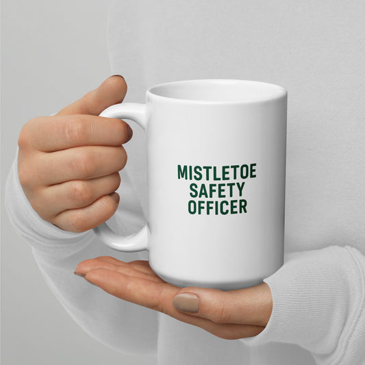 White mug with 'MISTLETOE SAFETY OFFICER' text held by a hand against a light gray background