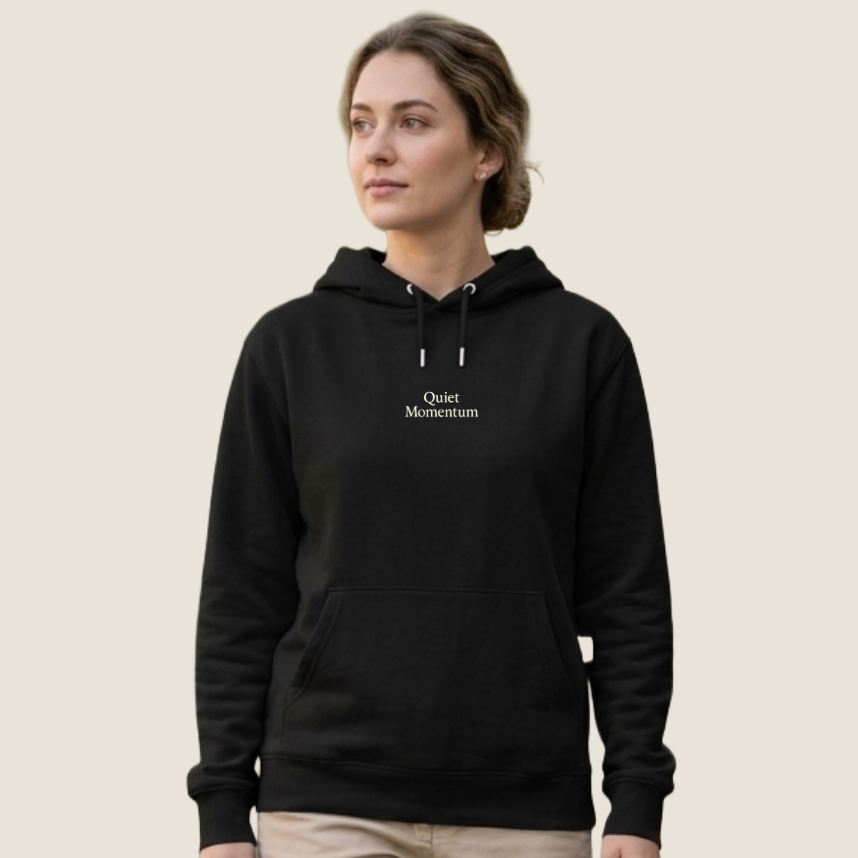 A woman wearing a black pullover hoodie with 'Quiet Motherhood' printed in white text on the chest, paired with beige pants, set against a solid light beige background.