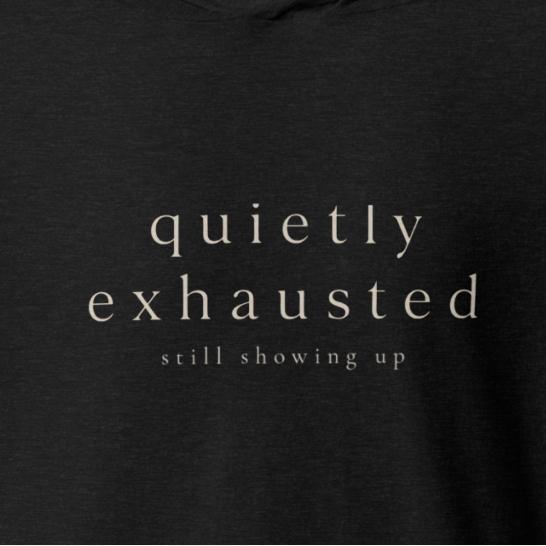 Close-up of a dark charcoal heathered t-shirt with the words 'quietly exhausted' and 'still showing up' printed in a light beige serif font.