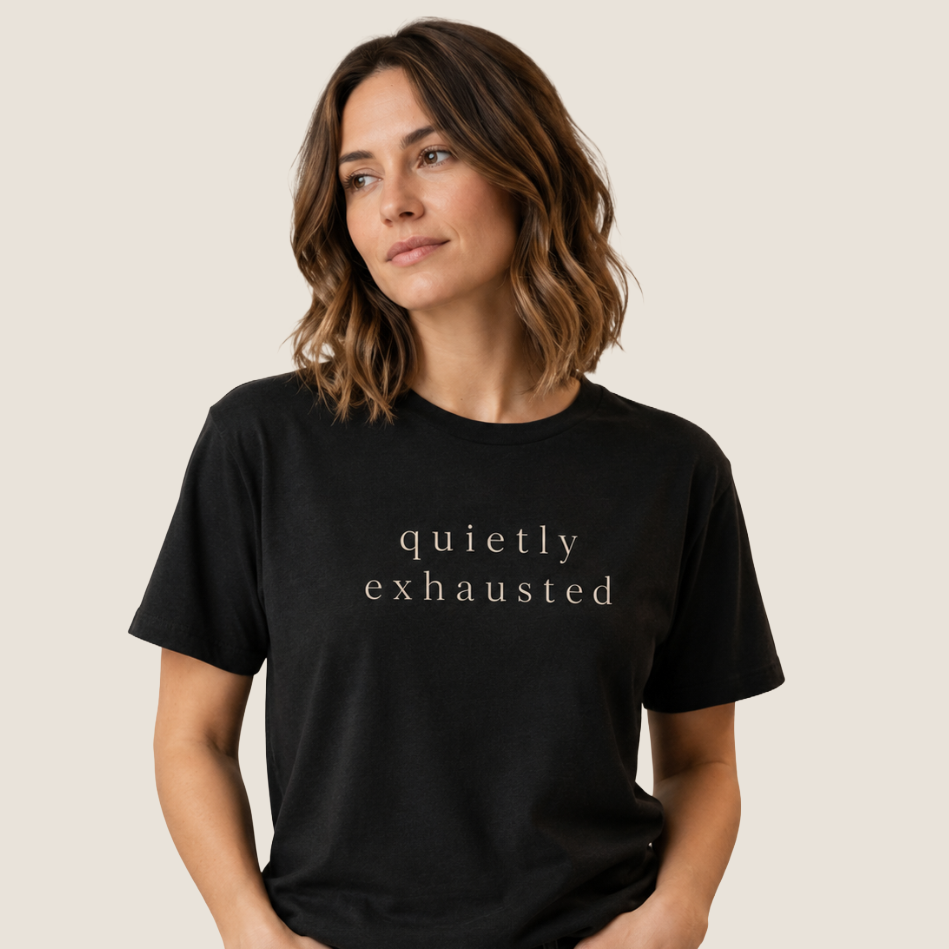 A woman with shoulder-length brown hair wearing a black crewneck t-shirt with the words 'quietly exhausted' printed in a light beige serif font, set against a solid light beige background.