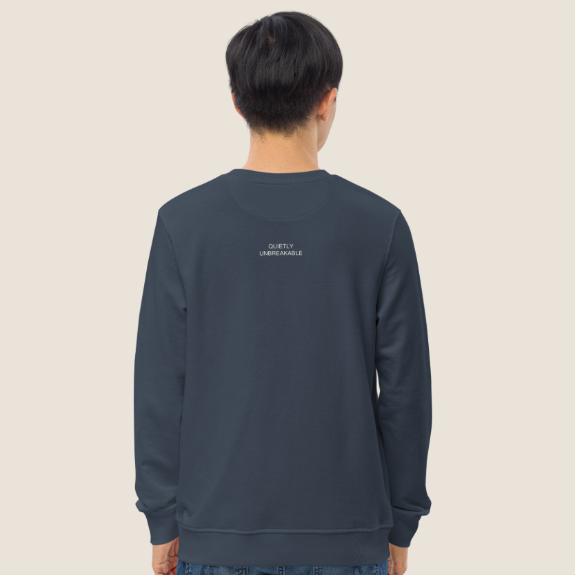 A person viewed from the back wearing a dark slate blue crewneck sweatshirt with small white text on the upper back, paired with blue jeans, against a solid light beige background.