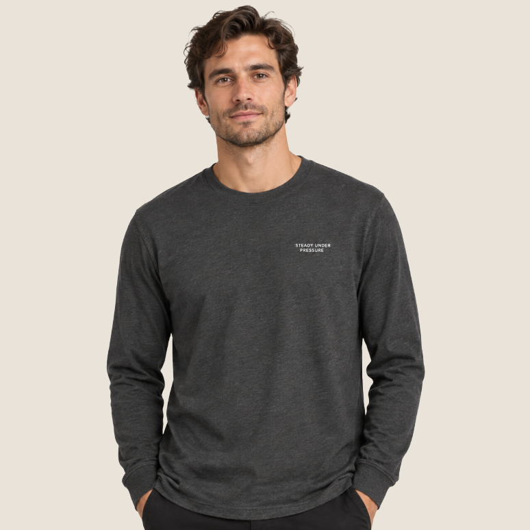 A man wearing a dark heather gray long-sleeve crewneck t-shirt with small white text on the left chest, standing against a solid light beige background.