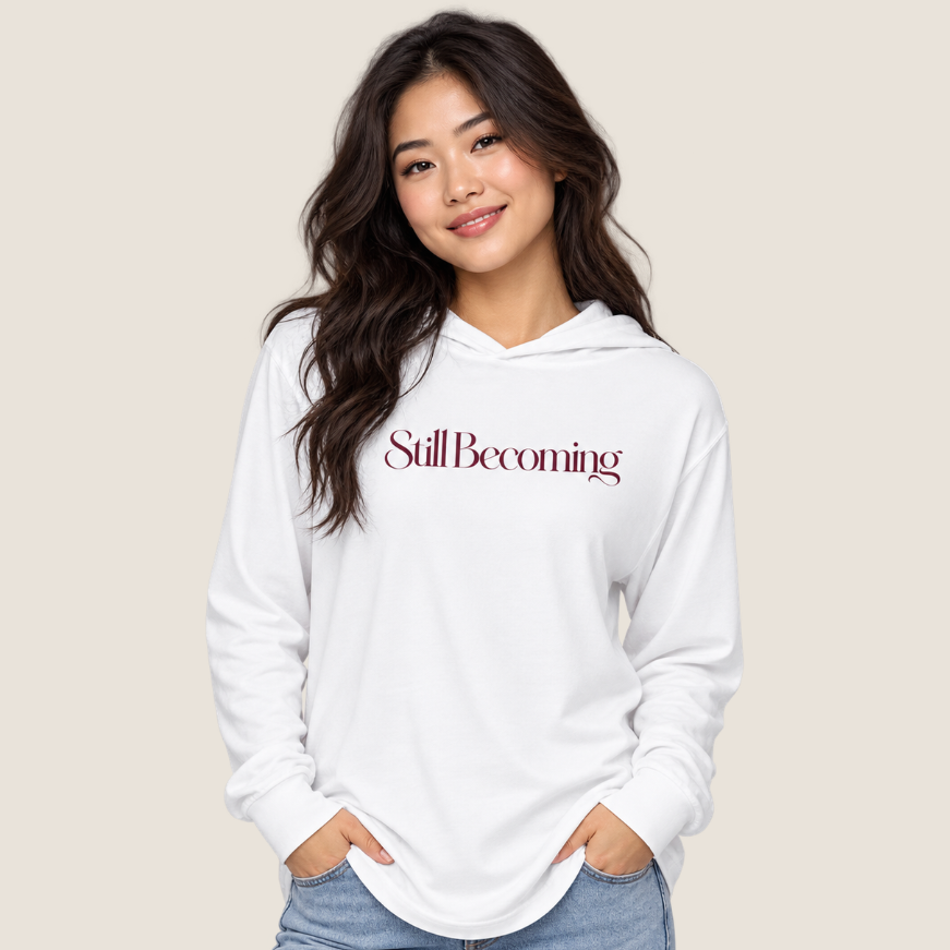 Woman wearing white hooded long sleeve tee with "Still Becoming" on the chest on a beige background.