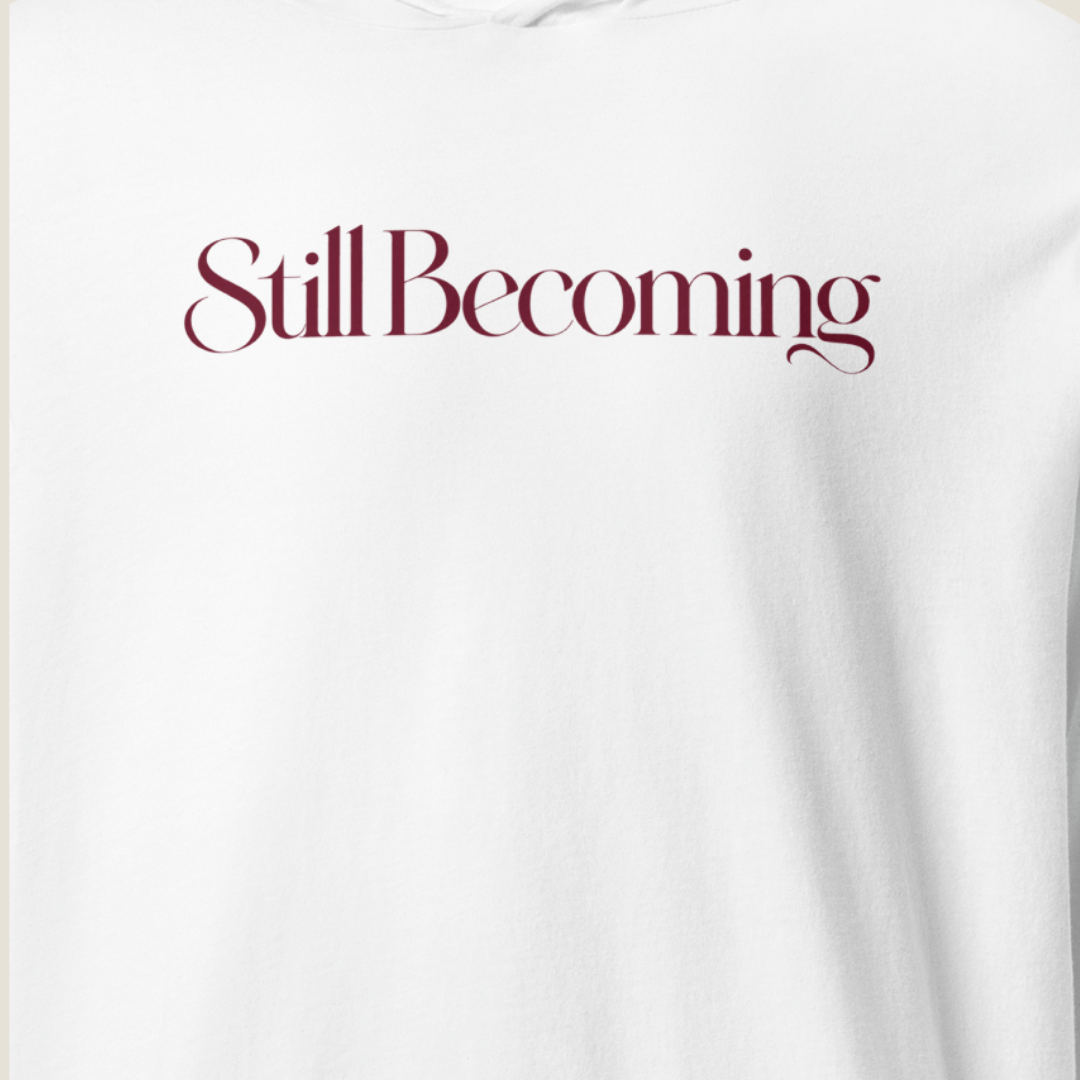 A close-up of a white sweatshirt featuring the phrase 'Still Becoming' printed across the chest in a dark burgundy serif font.