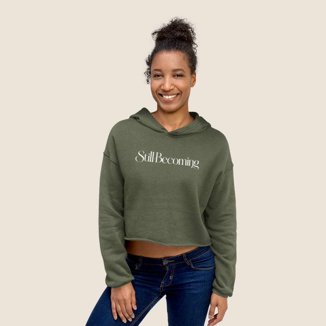 Woman wearing an olive cropped hoodie with "Still Becoming" text across text on a beige background.