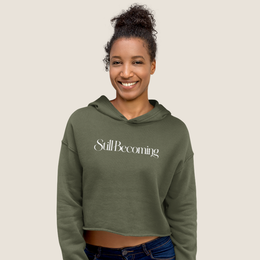 Woman wearing an olive cropped hoodie with "Still Becoming" text across text on a beige background.