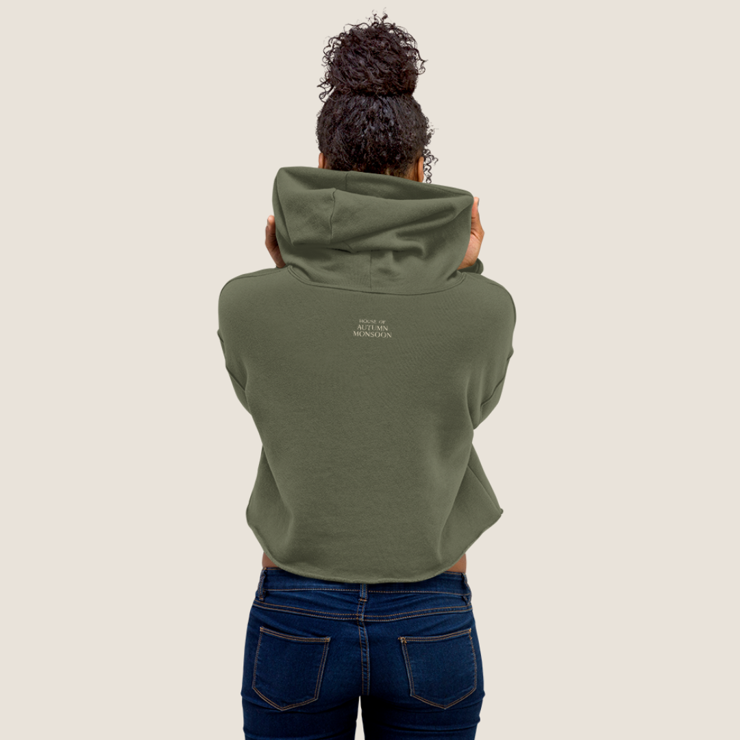 A person viewed from behind wearing an olive green cropped hoodie with small white text on the upper back, paired with dark blue jeans, against a plain light beige background.