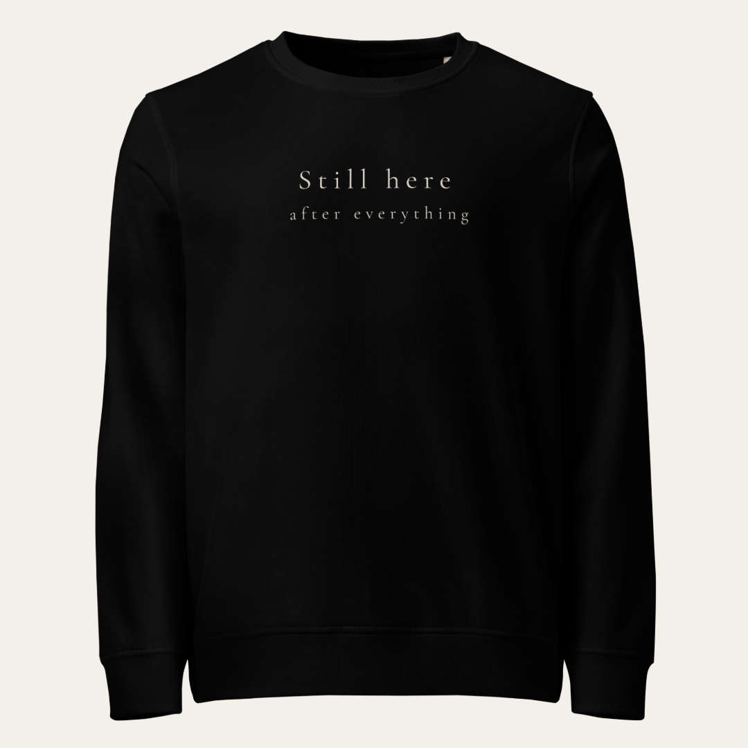 Black crewneck sweatshirt with Still here after everything on upper center chest.