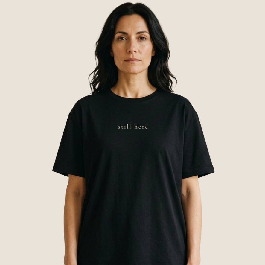 A woman wearing a black short-sleeve crewneck t-shirt with the text 'still here' printed in a small serif font on the chest, set against a solid light beige background.