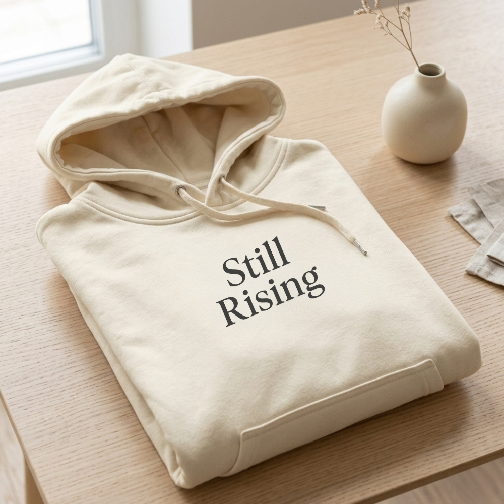 Beige hoodie with 'Still Rising' text on a wooden table, featuring the brand 'House of Autumn Monsoon'.
