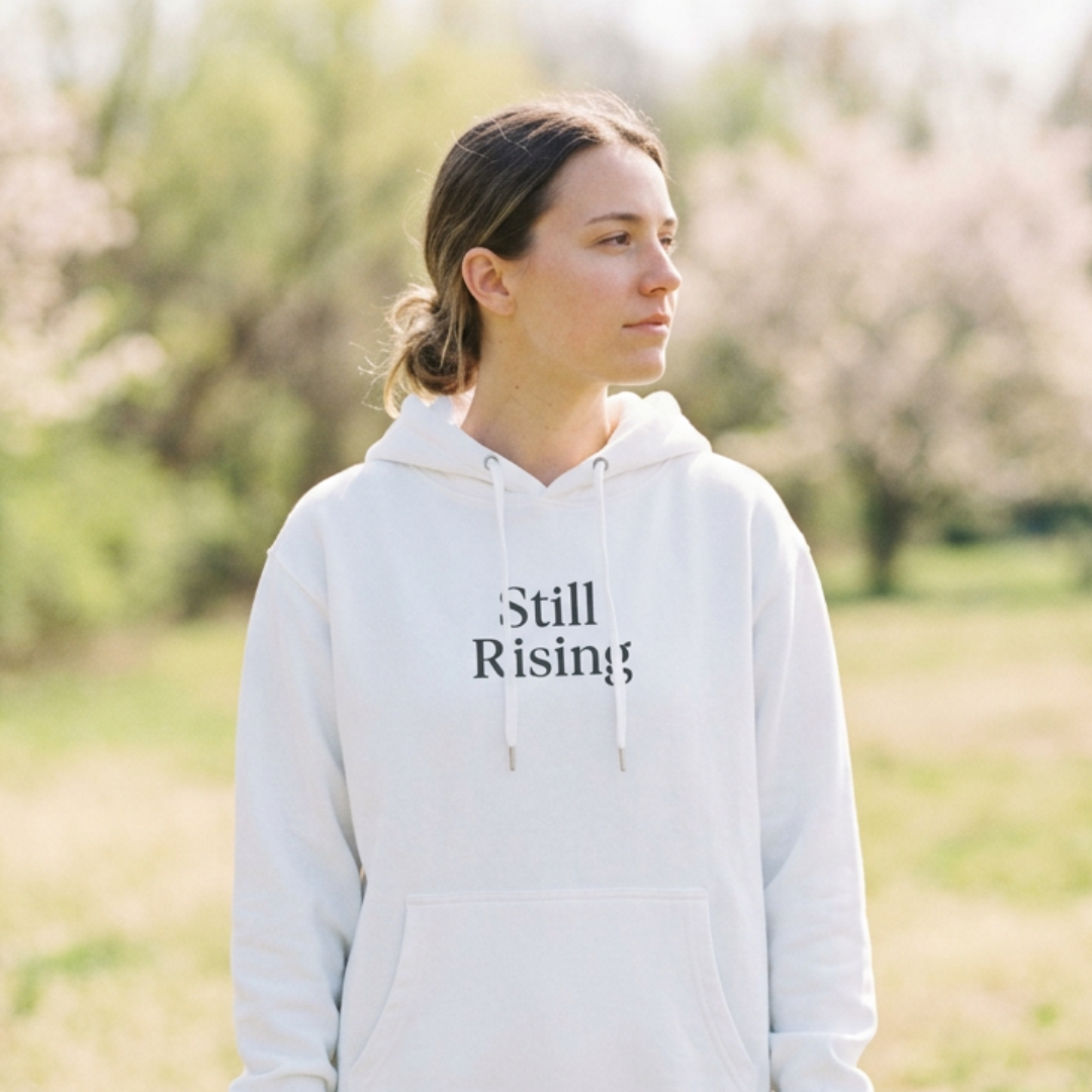 Female wearing the Still Rising white hoodie, looking to the side with a background of blooming trees.
