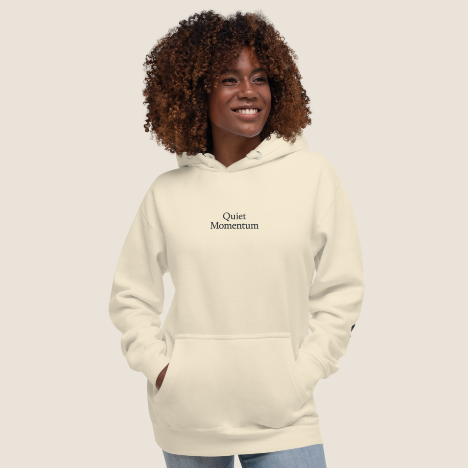A smiling woman with curly brown hair wearing a cream-colored pullover hoodie with the words 'Quiet Momentum' printed in black on the chest, paired with light blue jeans, set against a solid light gray background.