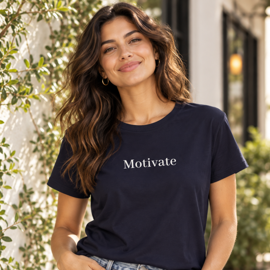 A woman wearing a navy blue crewneck t-shirt with the word 'Motivate' printed in white serif text across the chest, set against a softly blurred outdoor background with green foliage.