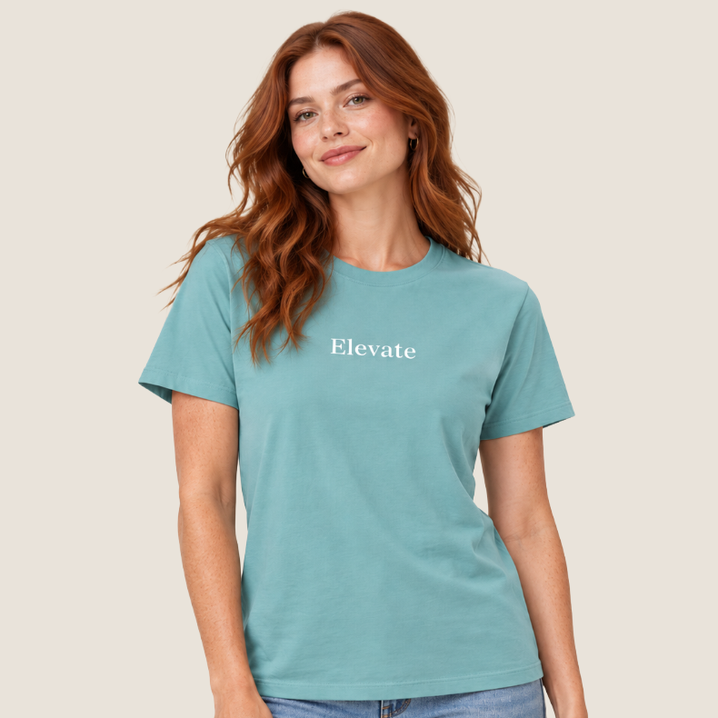 A woman modeling a short-sleeve crewneck t-shirt in a muted teal color with the word 'Elevate' printed in white serif text across the chest. She is wearing light-wash blue jeans against a solid, warm off-white studio background.