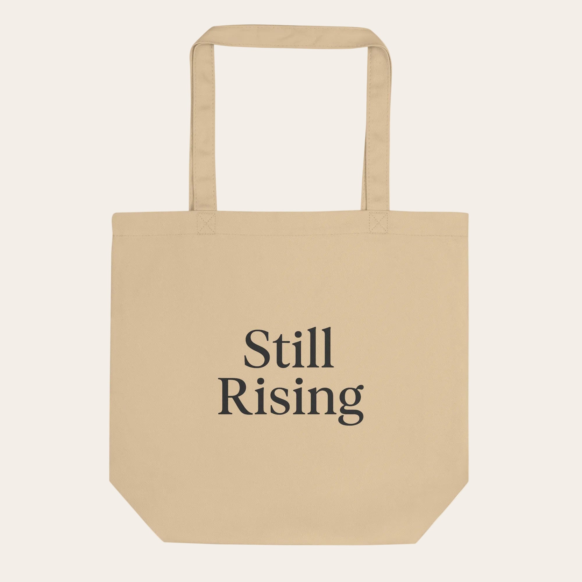 Beige tote bag with 'Still Rising' text on a white background