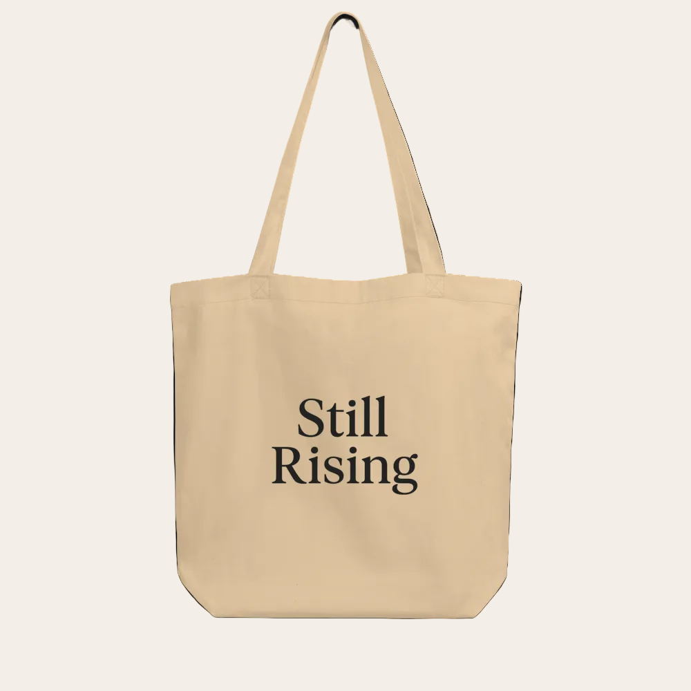 Beige tote bag with 'Still Rising' text on a white background