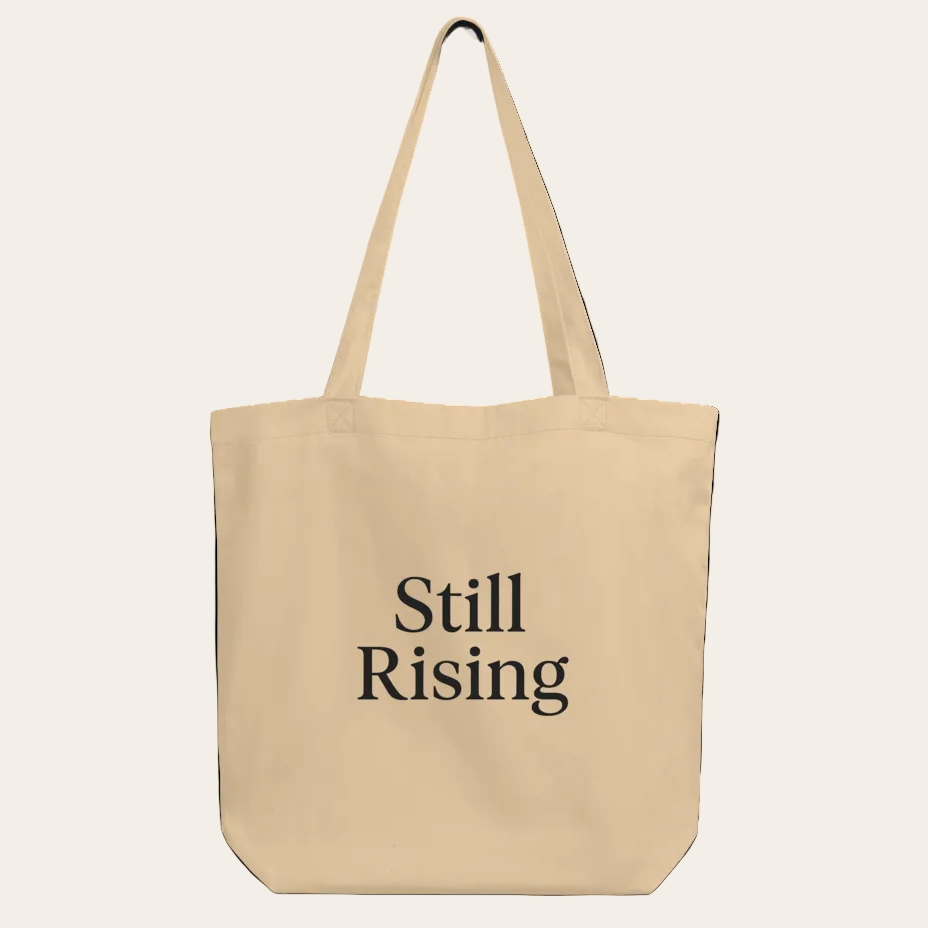 Beige tote bag with 'Still Rising' text on a white background