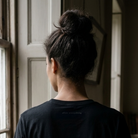 A person viewed from behind wearing a black crewneck t-shirt with the small white text 'after everything' printed on the back, standing in a dimly lit room near a window.