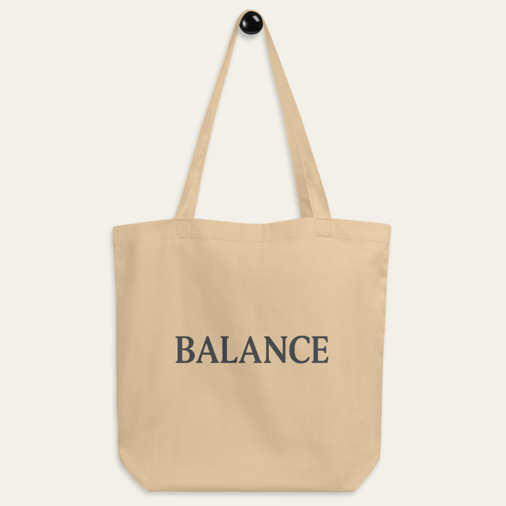 A beige canvas tote bag with the word 'BALANCE' printed in dark gray serif letters on the front, hanging from a black hook against a light off-white background.