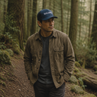 A man standing on a forest path wearing a navy blue baseball cap with 'BALANCE' embroidered in white, a brown overshirt over a dark gray t-shirt, and dark gray pants.