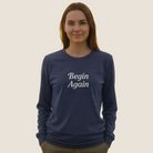 A woman wearing a navy blue long-sleeve crewneck sweatshirt with 'Begin Again' printed in white script on the chest.