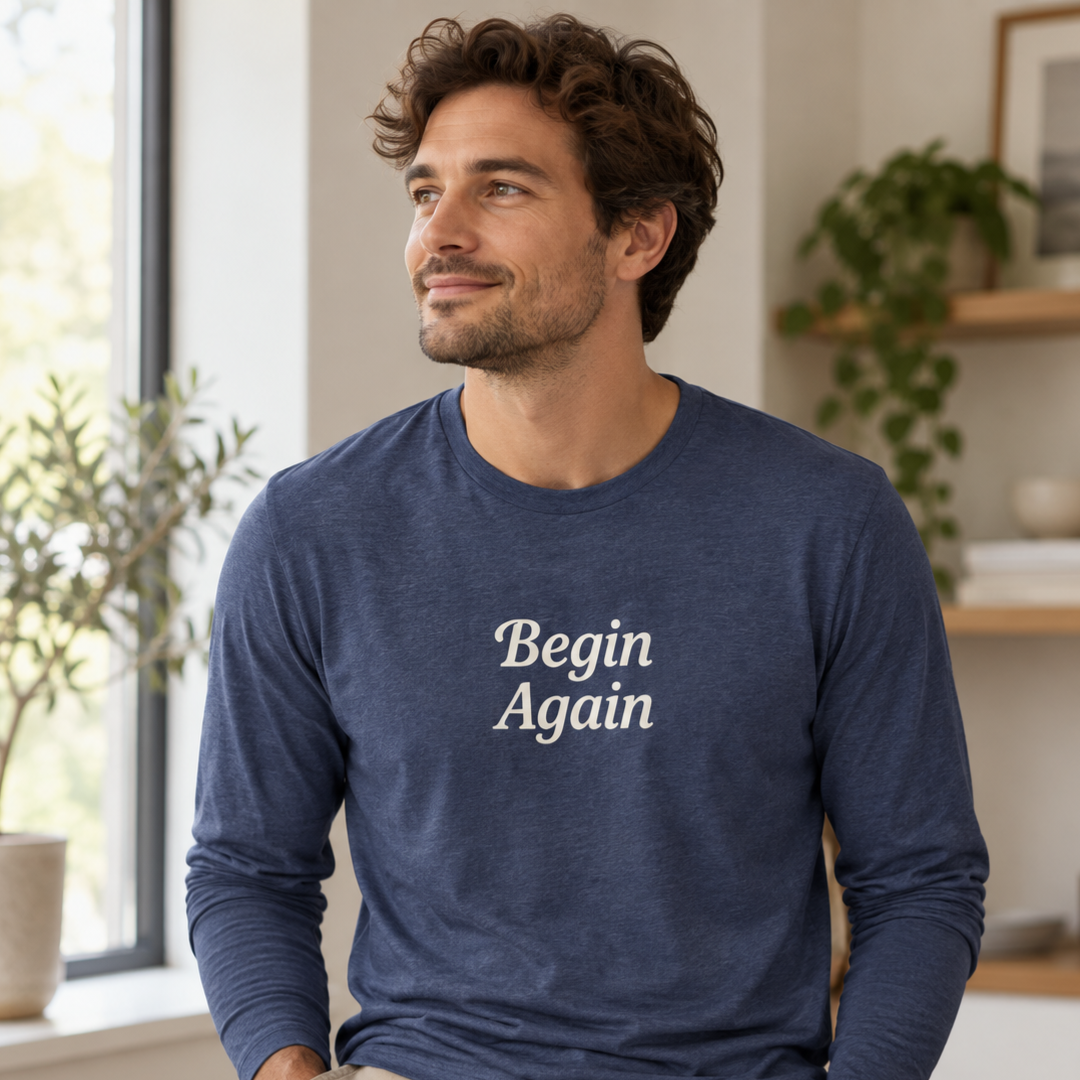 A man wearing a navy blue long-sleeve t-shirt with the words 'Begin Again' printed in white serif text on the chest, set against a softly blurred indoor background.