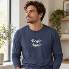 A man wearing a navy blue long-sleeve t-shirt with the words 'Begin Again' printed in white serif text on the chest, set against a softly blurred indoor background.