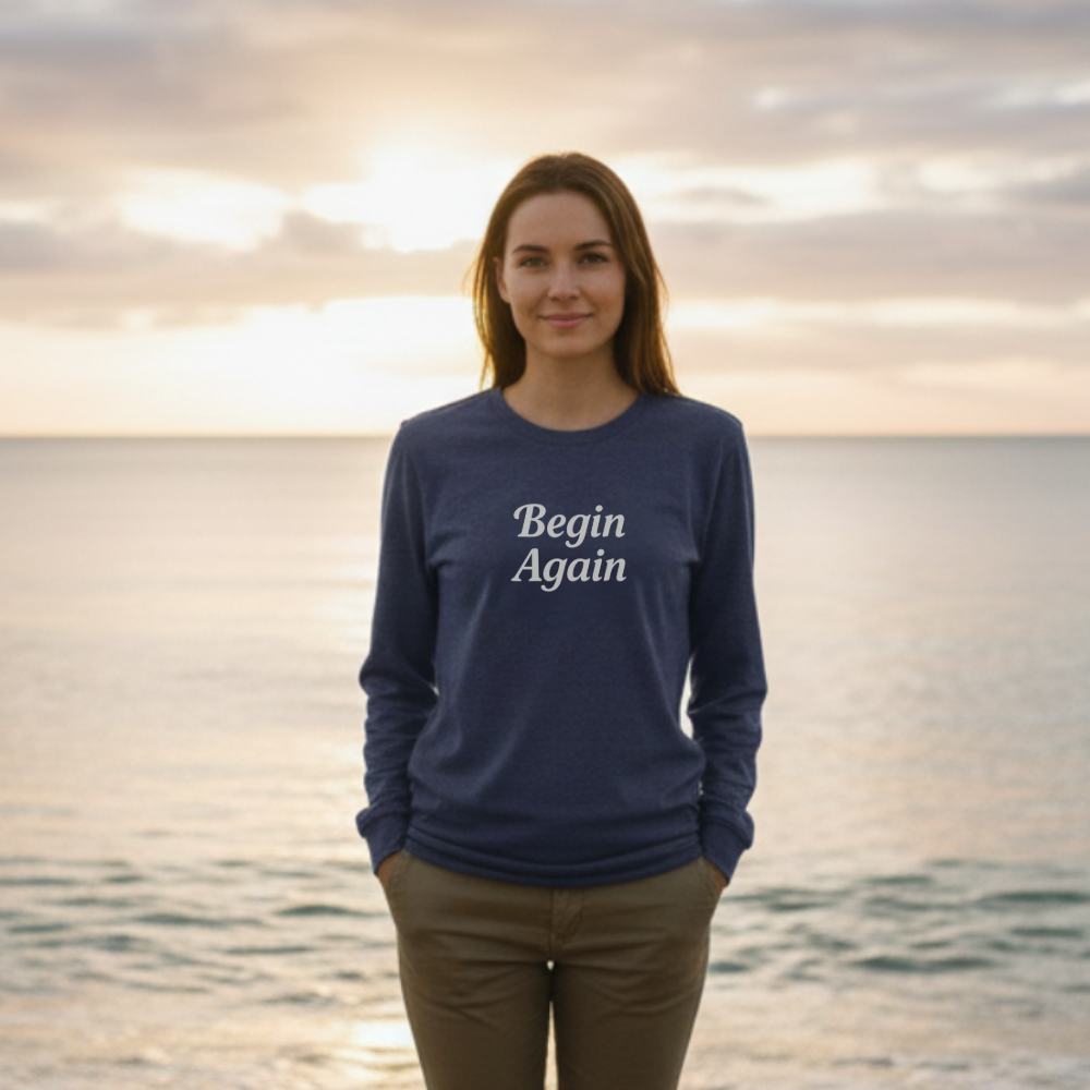 Person wearing a navy blue sweatshirt with 'Begin Again' text on a beach at sunset.