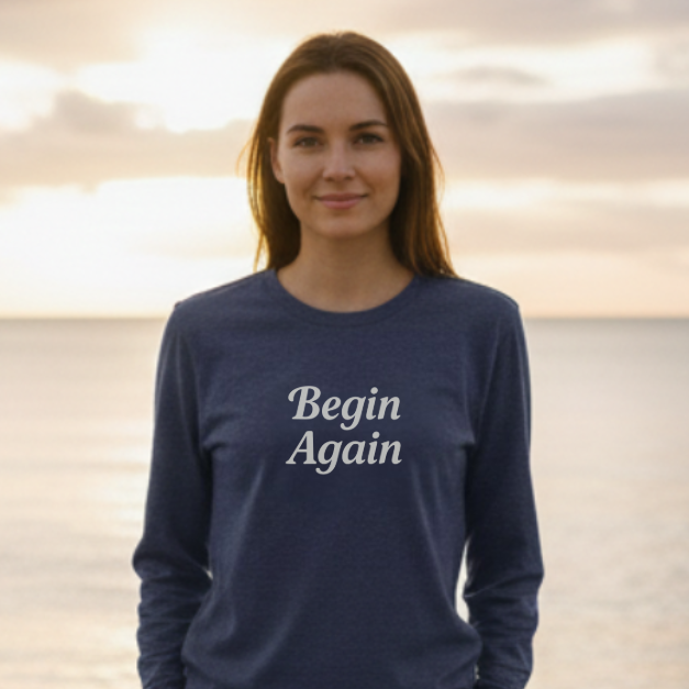 Person wearing a navy blue sweatshirt with 'Begin Again' text on a beach at sunset.