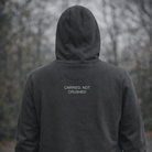 Person wearing a dark hoodie with 'CARRIED, NOT CRUSHED' text in a forest setting