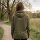 Female wearing a military green hoodie with Carried, Not Crushed in text on the back. She is standing on a path in the outdoors with trees ahead of her.