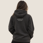 A woman viewed from behind wearing a dark charcoal gray hooded sweatshirt with the text 'CARRIED, NOT CRUSHED' printed in white on the back, set against a solid light beige background.