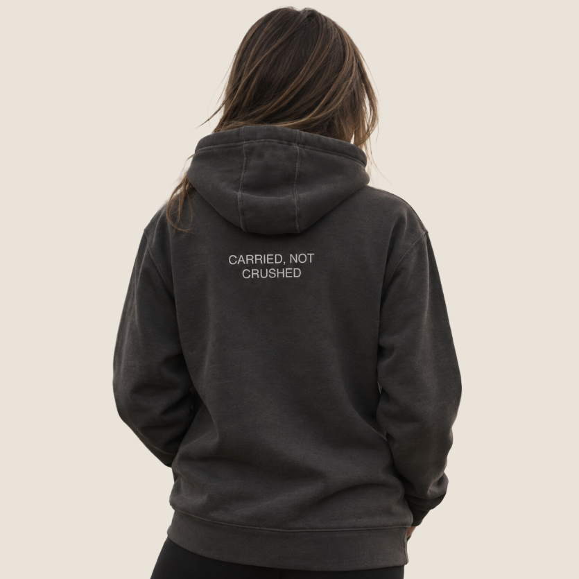 A woman viewed from behind wearing a dark charcoal gray hooded sweatshirt with the text 'CARRIED, NOT CRUSHED' printed in white on the back, set against a solid light beige background.