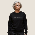 A woman with short gray hair wearing a black crewneck sweatshirt with the white printed text 'Depth Over Drama' on the chest, set against a solid light beige background.