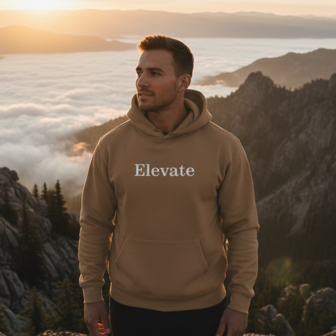 Man wearing a vintage brown hoodie with "Elevate" text on chest standing on a mountaintop with clouds below him.