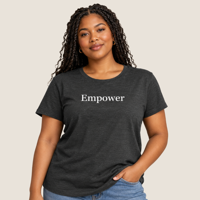 A woman with long black braids wearing a dark charcoal gray t-shirt with the word 'Empower' printed in white serif text across the chest, paired with light blue jeans, set against a solid light beige background.