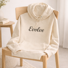 Beige hoodie with 'Evolve' text on a wooden chair in a minimalistic room.