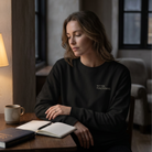 Woman sitting at a table with a notebook and pen, wearing a black sweatshirt with text.