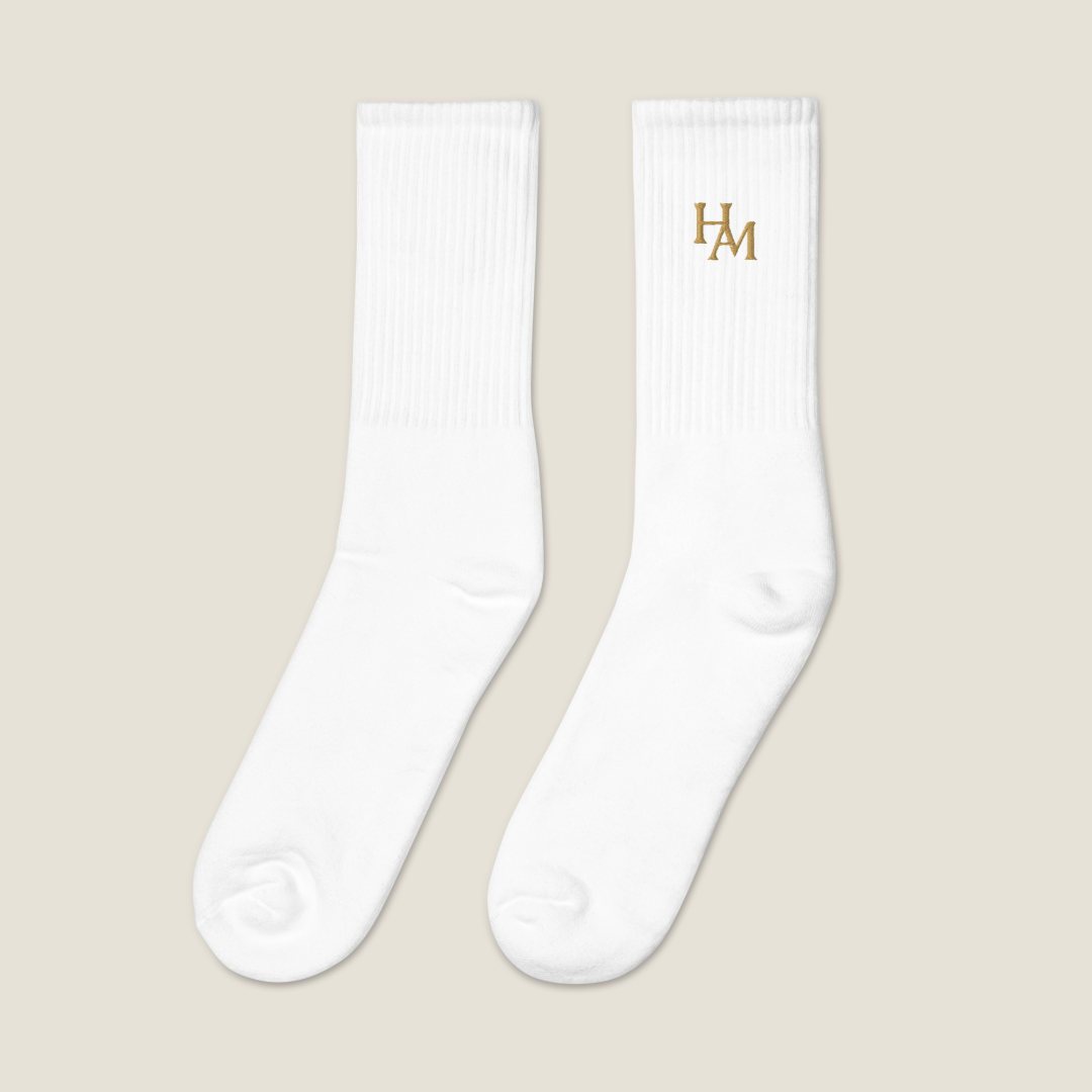 A pair of white crew socks with a gold embroidered 'HAM' logo on the upper right sock, displayed against a solid light beige background.