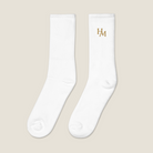 A pair of white crew socks with a gold embroidered 'HAM' logo on the upper right sock, displayed against a solid light beige background.